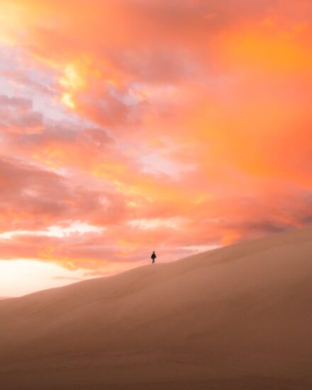 unrecognizable tourist walking along hilly desert under sunset sky